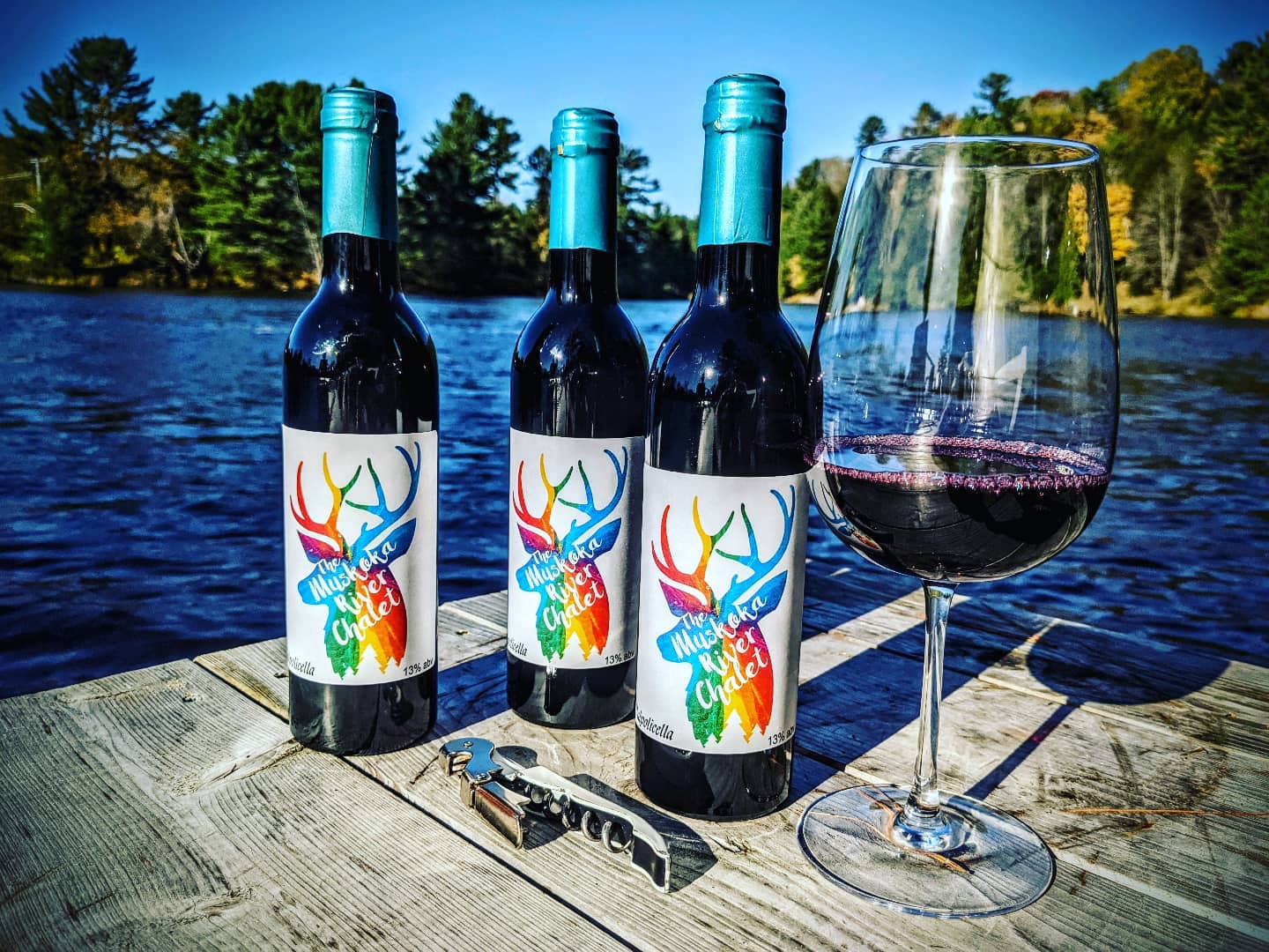 The Muskoka River Chalet branded wine bottles with a glass of red wine on the dock overlooking the Muskoka River