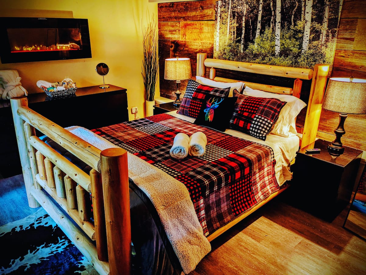 The King's Den bedroom at The Muskoka River Chalet featuring a log-frame king bed, plaid bedding, wall-mounted electric fireplace, and reclaimed wood accent wall